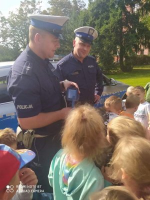 policjanci na placu przed przedszkolem stoja obok radiowozu, pokazują dzieciom sprzęt policyjny dzieci stoją tyłem d patrzą na policjanta