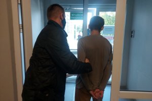 Korytarz, drzwi wyjściowe, policjant w ubraniu cywilnym stoi tyłem, trzyma pod rękę zatrzymanego mężczyznę,który ręce ma skute kajdankami . wychodzą przez drzwi na plac