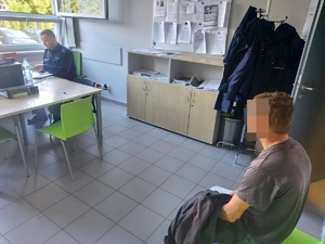 zatrzymani za kradzież, siedzą na krześle w pokoju, w tle stoi policjant w mundurze , Przed nimi okno , widać także umeblowanie pokoju. szafki w  kolorze jasnym, na biurku komputer